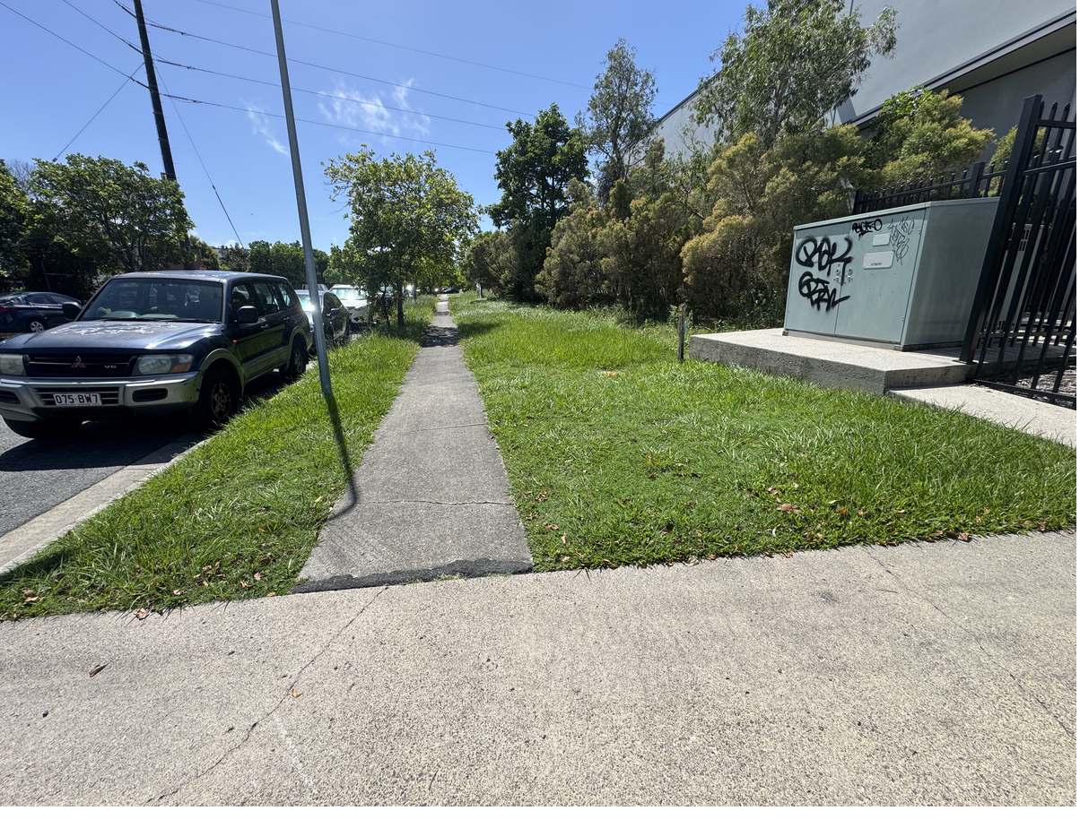 Overgrown grass verge and footpath along an industrial estate frontage at Warana before scheduled grounds maintenance
